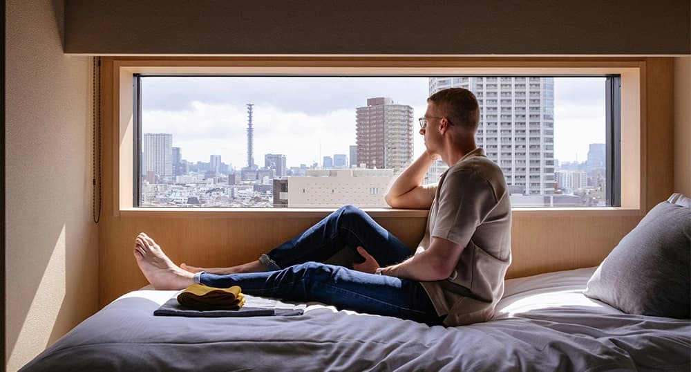 Man relaxes on bed overlooking window view of Japanese city.