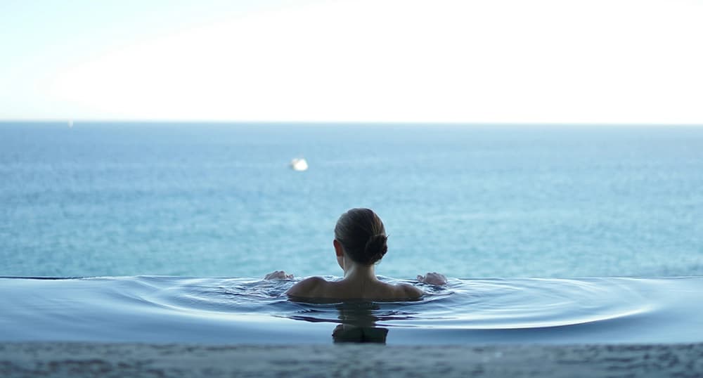 Woman on wellness retreat stares out over the ocean in a plunge pool.