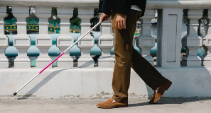 Visually impaired man walks on city sidewalk with white cane.