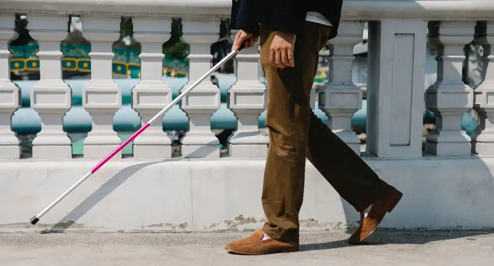 Visually impaired man walks on city sidewalk with white cane.
