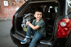 Smiling young boy sits in the trunk area of a black SUV packed for a trip.