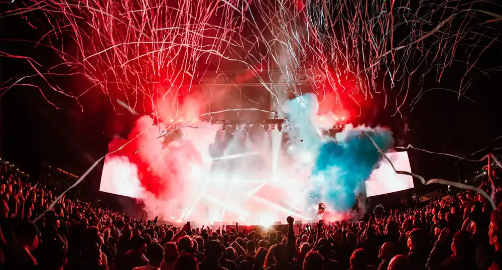 Colorful fireworks and lights over a stage with large crowd of people.