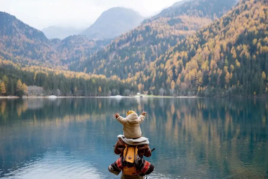 Toddler sits on parent's shoulders as both overlook scenic view of a lake and mountains.