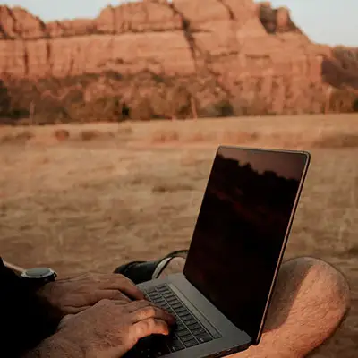 Man types on a laptop outdoors with red rock cliffs in the background.