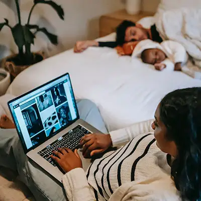 Woman works on a laptop as father and baby nap on a bed nearby.